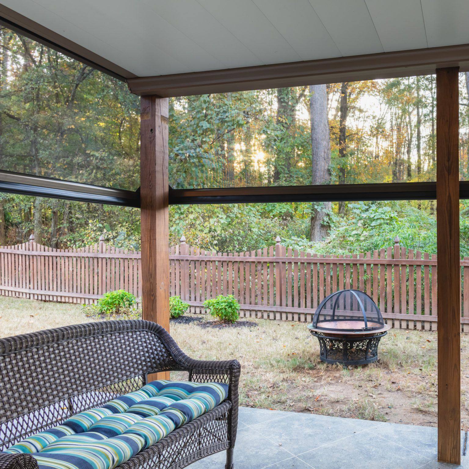Sunlight filtering through outdoor screens beside wicker seating and a wooded garden.